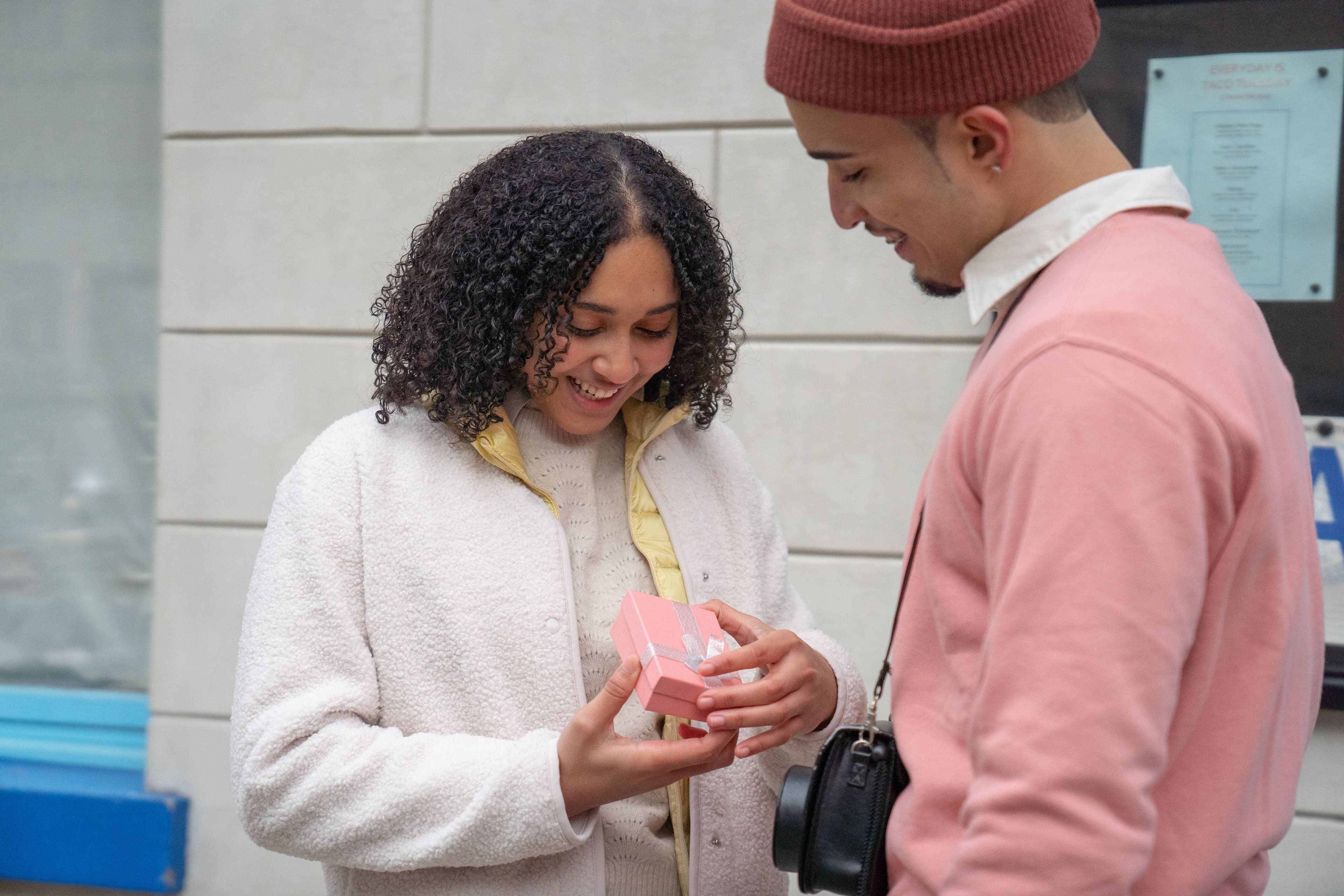 A woman smiles as she hands a wrapped gift box to her boyfriend on a sunny city street